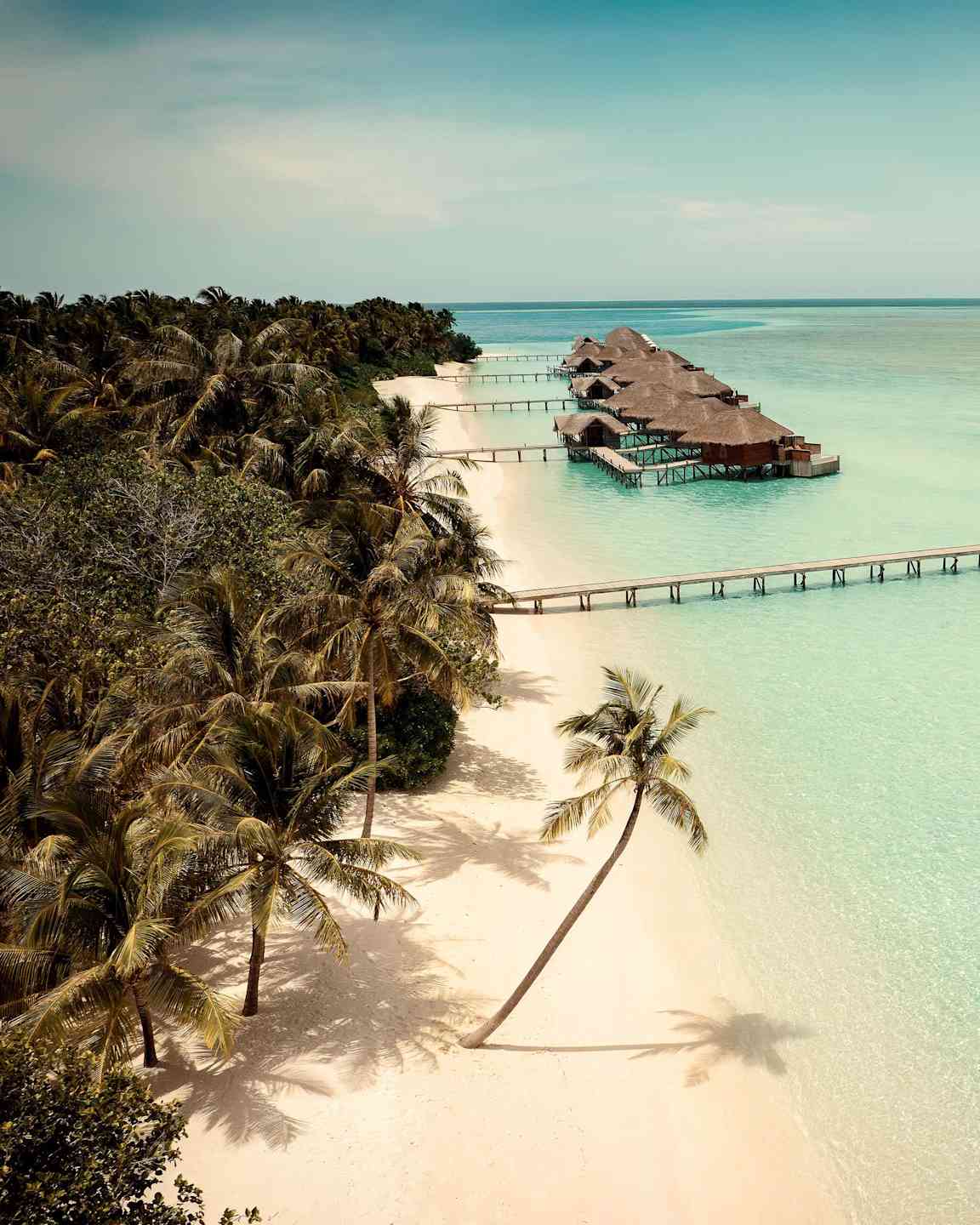 En vit sandstrand med palmer vid sidan av en turkos lagun där flera bungalows på pålar är placerade. (AI-genererad text)