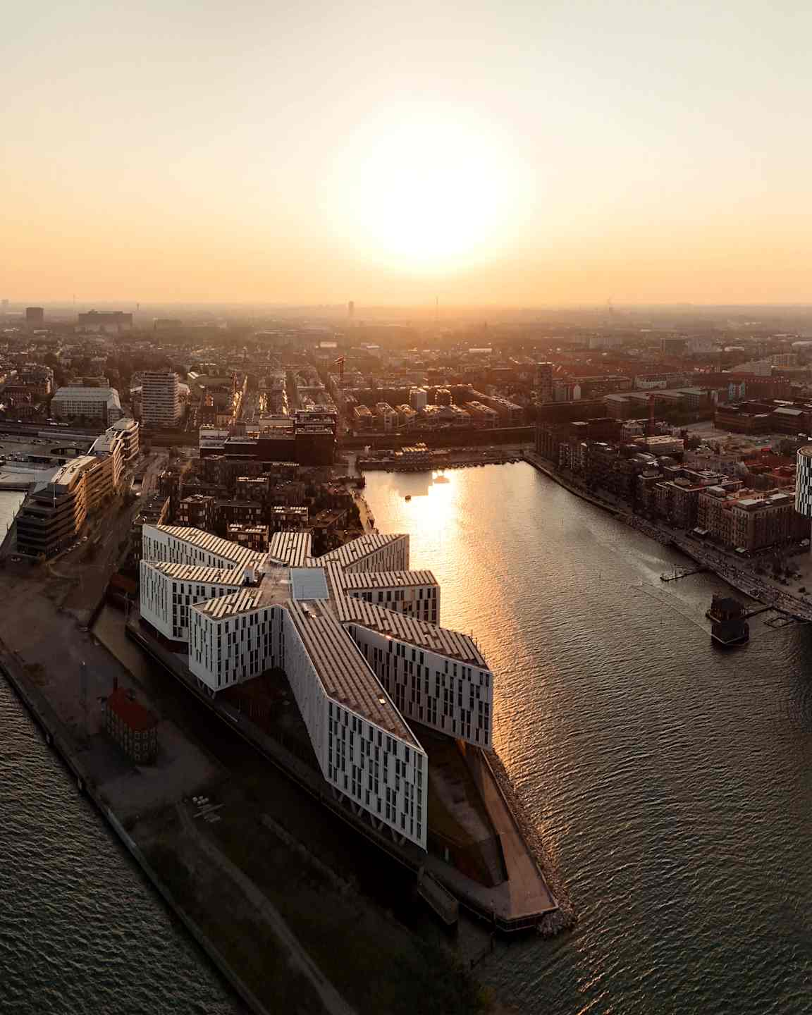 Flygbild över en stad med moderna byggnader vid vattnet och en solnedgång i bakgrunden. (AI-genererad text)