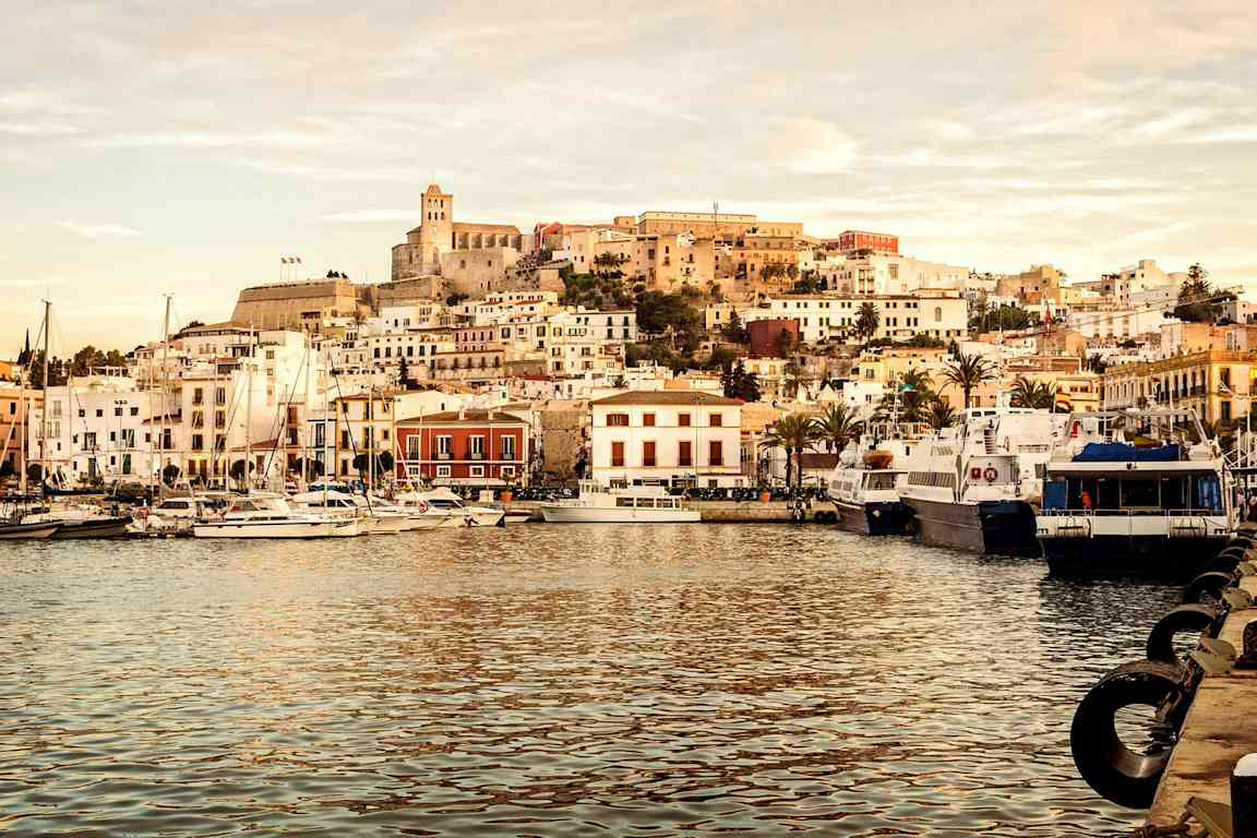 Vy över hamnen med segelbåtar och byggnader, samt en kulle med äldre arkitektur i bakgrunden. (AI-genererad text)