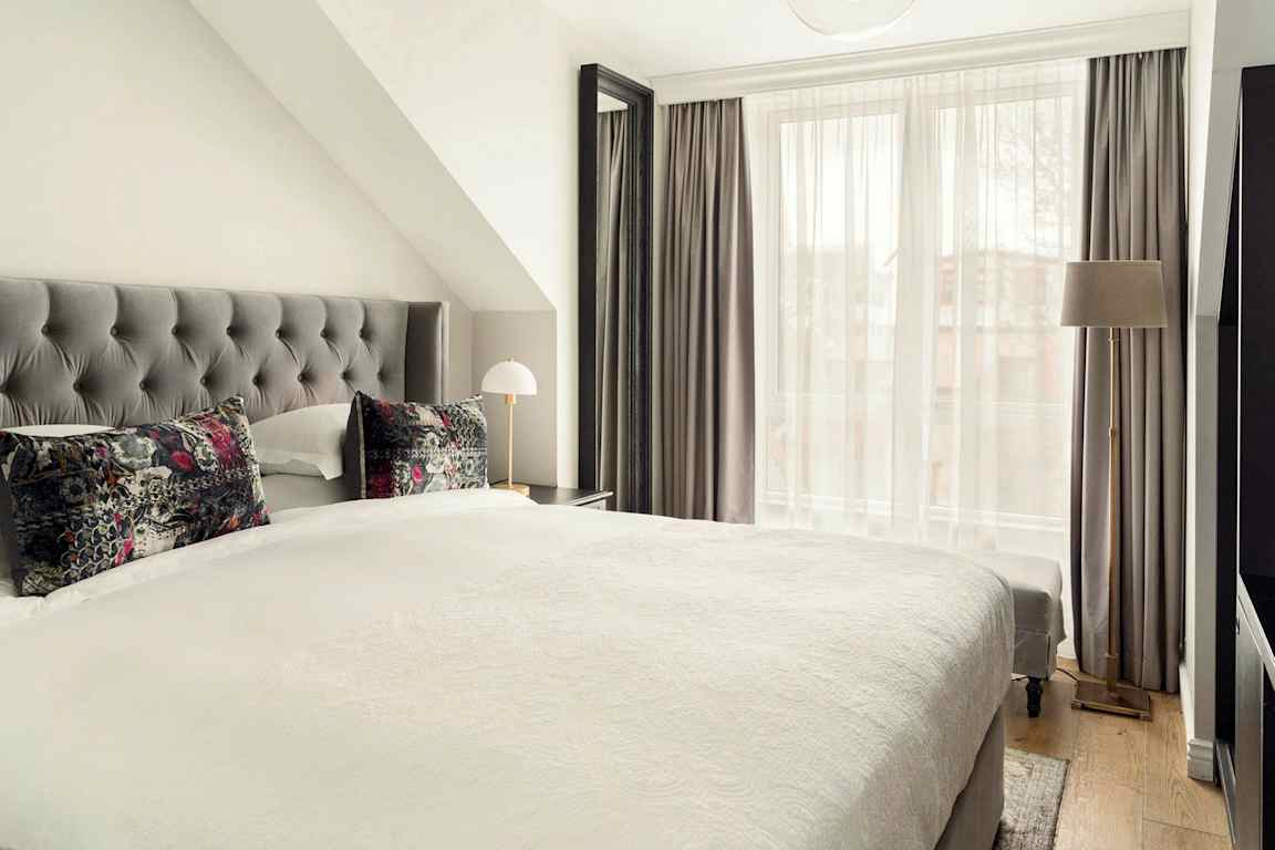 Ett hotellrum med en stor säng, grå sänggavel och färgglada kuddar. Gardiner täcker ett stort fönster. (AI-genererad text)