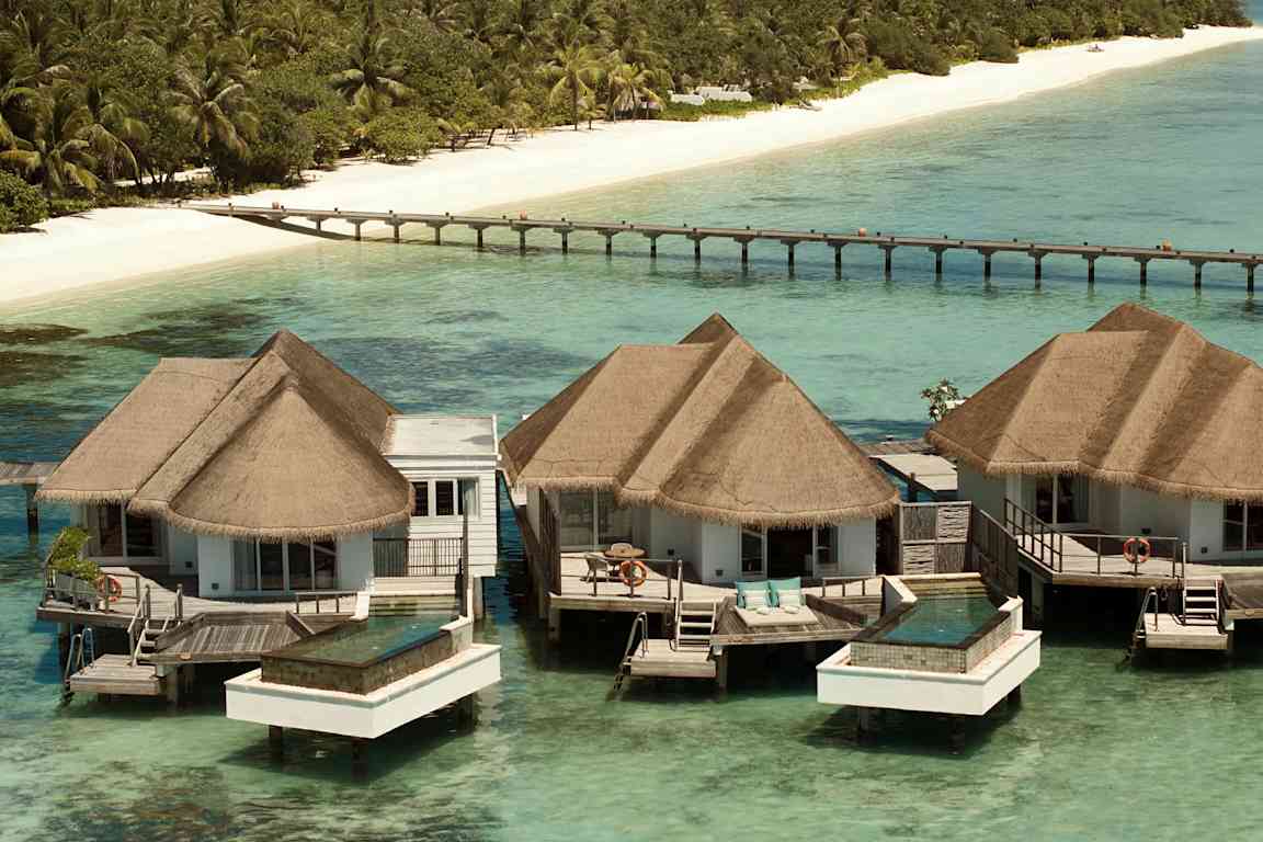 Bilden visar bungalows med halmtak belägna på vattnet, en lång brygga och en vit sandstrand med grönska i bakgrunden. (AI-genererad text)