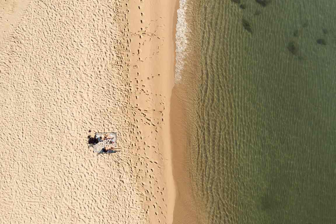 Flygbild av en strand med sand, vatten och personer liggande på en handduk. (AI-genererad text)
