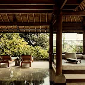 Interiören i en öppen tropisk träbyggnad med utsikt mot grönska, en hörnsoffa och två solstolar. (AI-genererad text)