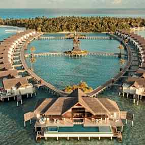 Flygfoto över bungalower som står på pelare i havet och formar ett hjärta mot en grönskande ö i bakgrunden. (AI-genererad text)