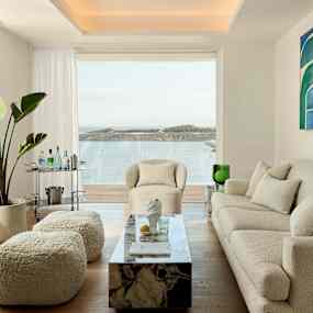 Ett modernt vardagsrum med beige möbler och konstverk på väggen, stora fönster visar en havsutsikt. (AI-genererad text)