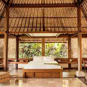 Ett rymligt sovrum med trädetaljer och stora fönster som vetter mot en grönskande trädgård. En säng är centralt placerad med omgivande möbler. (AI-genererad text)