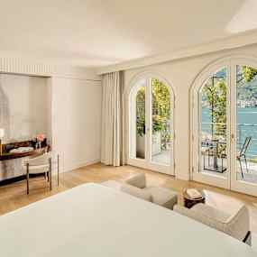 Lyxigt hotellrum med stora välvda fönster som öppnar upp mot en balkong med utsikt över en sjö. (AI-genererad text)