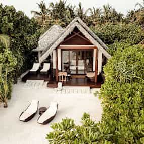 En tropisk villa med halmtak vid stranden, omgiven av grönska och palmer. (AI-genererad text)