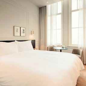 Ett hotellrum med en stor säng, ett fönster med gardiner och ett litet bord med två stolar. (AI-genererad text)