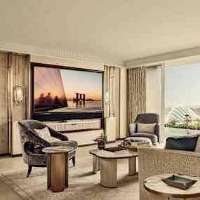 Ett elegant vardagsrum med moderna möbler, inklusive soffor och fotöljer, och en stor TV på väggen. Ett fönster visar utsikt över en trädgård och stadens silhuett. (AI-genererad text)