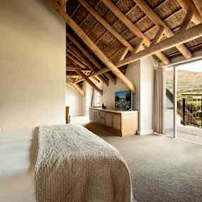 Ett sovrum med en dubbelsäng, en TV på en möbel, och en balkong med utsikt över en grönskande dal. (AI-genererad text)