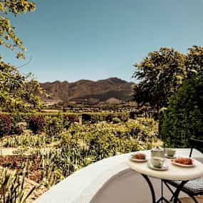 En liten cafébord med te och croissanter står i förgrunden, med utsikt över en grönskande trädgård och berg i bakgrunden. (AI-genererad text)