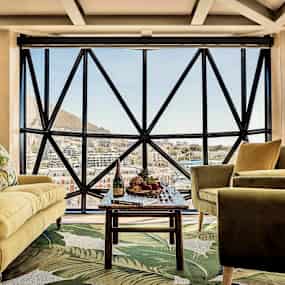 Ett vardagsrum med stora fönster och utsikt över en stad samt berg. Rummet har en gul soffa, gröna fåtöljer och ett bord med frukt. (AI-genererad text)