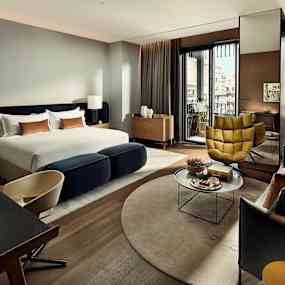 Ett modernt hotellrum med stor säng, sittgrupp och utsikt mot byggnader. (AI-genererad text)