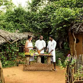 Personer i kökskläder står vid ett bord med grönsaker och råvaror i en lantlig miljö med uppbyggda hyddor. (AI-genererad text)