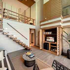 Ett modernt vardagsrum med högt i tak, trägolv och en trappa som leder till en övre nivå. Det finns en hörnmöbel med böcker och en TV, samt en inomhusväxt. (AI-genererad text)