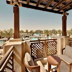 En terrass med trästolar och bord, med utsikt över poolområde och palmer. (AI-genererad text)