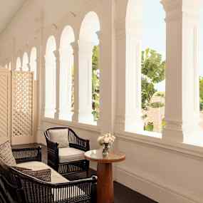 Ett ljust rum med svarta rottingmöbler och ett litet bord. Bakgrunden består av vitmålade valv och gröna trädkronor. (AI-genererad text)