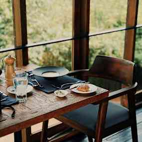 Ett dukat bord med en tallrik soppa, ett glas vatten och en brödservering, placerat vid ett fönster med utsikt mot grönska. (AI-genererad text)