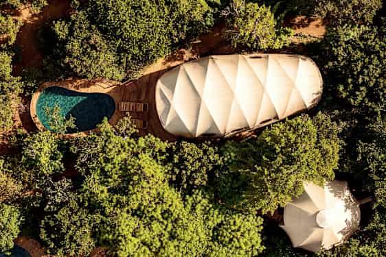 Flygfoto över en tältliknande byggnad, pool med trädäck och omgivande grönska. (AI-genererad text)