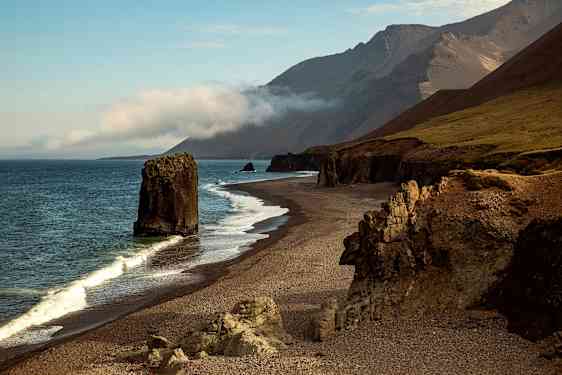 Bild av en klippig kust med en stor klippa i vattnet och berg i bakgrunden. (AI-genererad text)