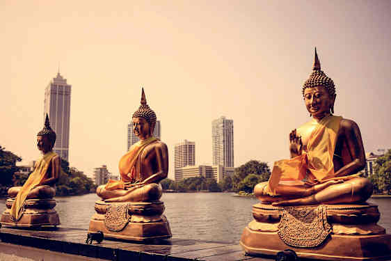 Tre gyllene buddhastatyer vid en vattendrag med moderna byggnader i bakgrunden. (AI-genererad text)