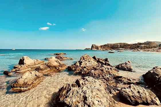 En strand med klippformationer och klart vatten intill en bergssluttning. (AI-genererad text)