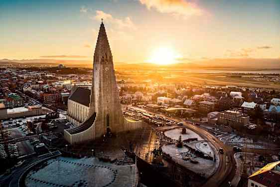 En kyrka med modern arkitektur och en hög tornstruktur, omgiven av en stad och ett landskap under en solnedgång. (AI-genererad text)