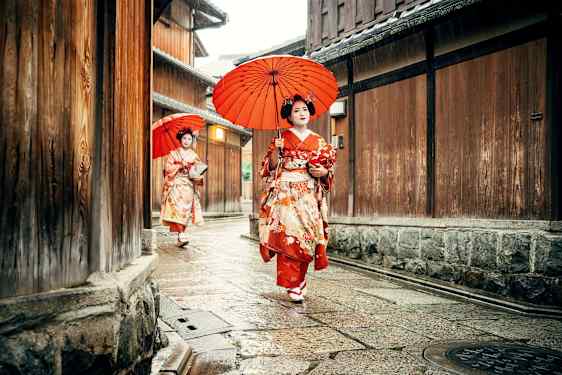 Två personer klädda i traditionell japansk kimono med röda paraplyer går längs en smal gata omgiven av trähus. (AI-genererad text)