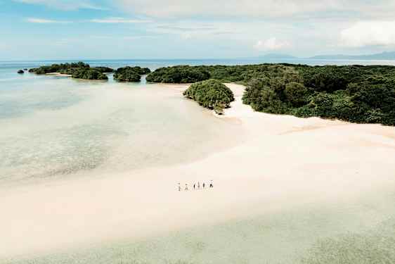 En flygbild av en sandstrand med gröna buskar och omgivande små öar i ett klart, blått hav. En grupp människor går längs stranden. (AI-genererad text)