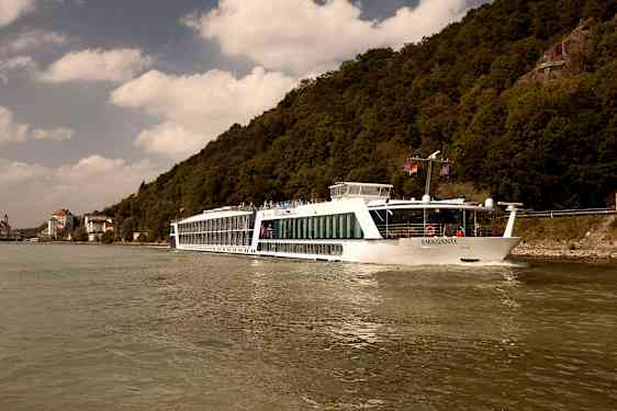 Ett fartyg, märkt med namnet 'AMADANTE', seglar på en flod nära skogsklädda berg och byggnader. (AI-genererad text)