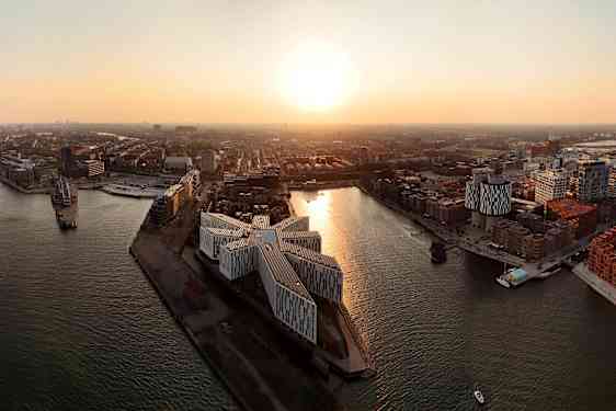 Flygbild över en stad med moderna byggnader vid vattnet och en solnedgång i bakgrunden. (AI-genererad text)