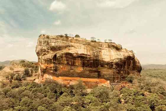 Bild av en stor klippa omgiven av skog under en blå himmel med vita moln. (AI-genererad text)