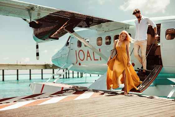 En kvinna och en man stiger av ett vattenflygplan som står vid en brygga över klart, blått vatten. Kvinnan bär en gul klänning. (AI-genererad text)