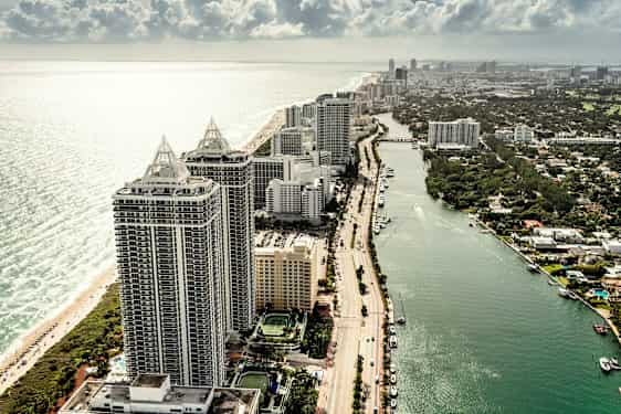 Flygvy över höga byggnader vid stranden, bredvid en kanal med båtar och palmträd. (AI-genererad text)