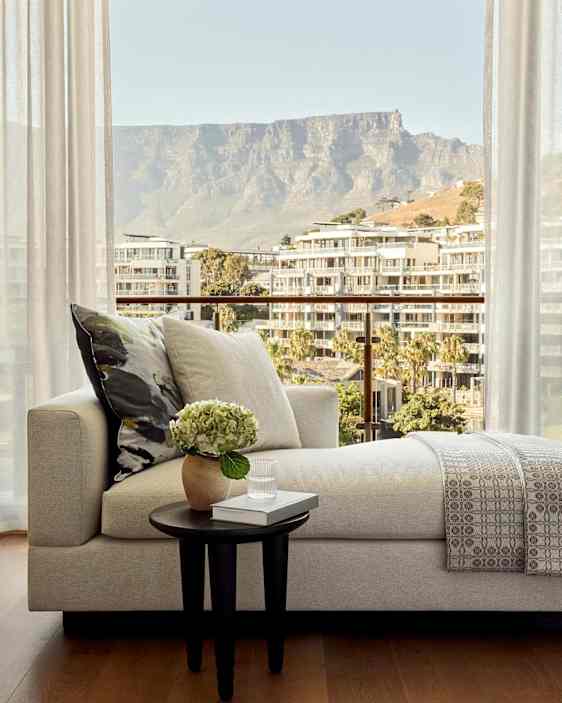 Ett vardagsrum med soffa, bord och utsikt över byggnader och ett berg genom stora fönster. (AI-genererad text)