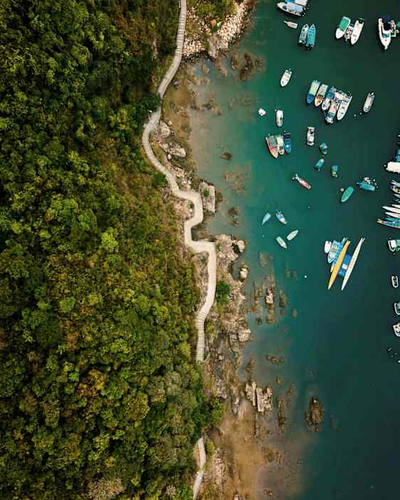 Luftvy över en kurvig stig längs en grön skog nära en klippig kust med båtar på vattnet. (AI-genererad text)