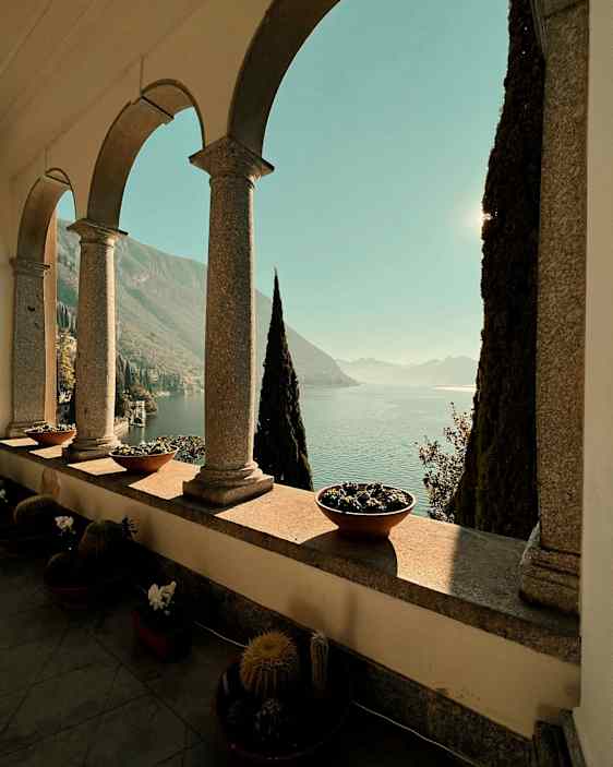 Bildens motiv består av en terrass med valvbågar, blomkrukor och kaktusar, med utsikt över en sjö och berg i bakgrunden. (AI-genererad text)
