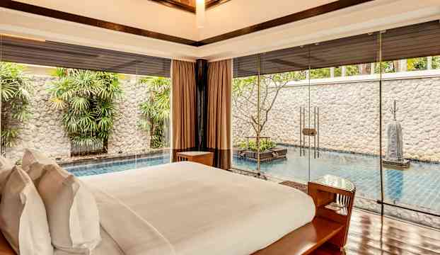 Ett sovrum med en stor säng och utsikt över en privat pool och grönskande omgivningar genom glasväggar. (AI-genererad text)