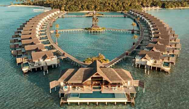 Flygfoto över bungalower som står på pelare i havet och formar ett hjärta mot en grönskande ö i bakgrunden. (AI-genererad text)