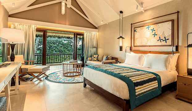 Hotellrum med dubbelsäng, sittgrupp och utsikt över grönskande natur. (AI-genererad text)