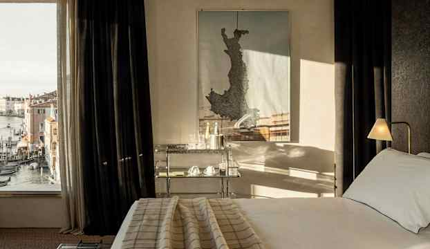 Ett hotellrum med en säng, ett nattduksbord och en karta på väggen, samt ett fönster med utsikt över gondoler och byggnader. (AI-genererad text)
