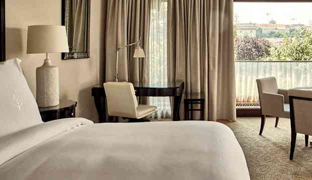 Ett hotellrum med en bäddad säng, ett skrivbord med en skrivbordsstol samt två fåtöljer runt ett litet bord. I bakgrunden finns ett fönster med gardiner och utsikt. (AI-genererad text)