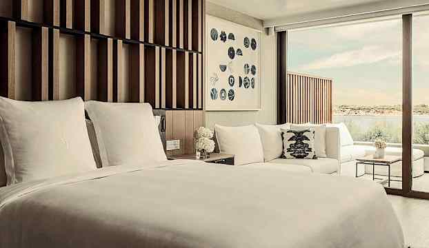 Ett hotellrum med en stor säng, soffa och fönster som vetter mot vatten. (AI-genererad text)