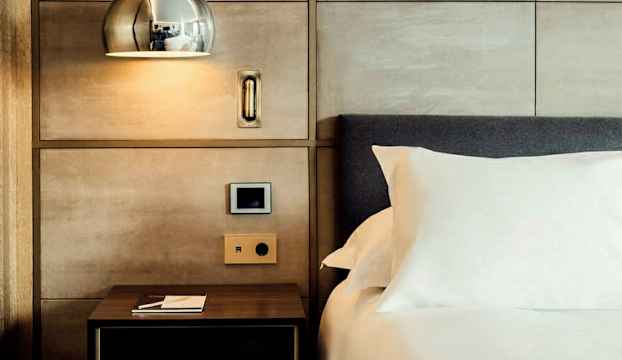 Närbild på en uppbäddad säng med en vit kudde och sidobord under en vägglampa. (AI-genererad text)