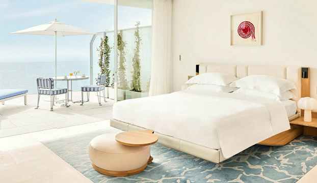 Ett ljust hotellrum med en dubbelsäng och möbler bredvid ett öppet område som leder till en terrass med utsikt mot havet. (AI-genererad text)