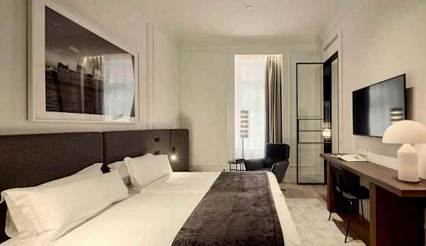 Ett hotellrum med en stor säng, ett skrivbord med en lampa, en TV på väggen och en fåtölj vid fönstret. (AI-genererad text)