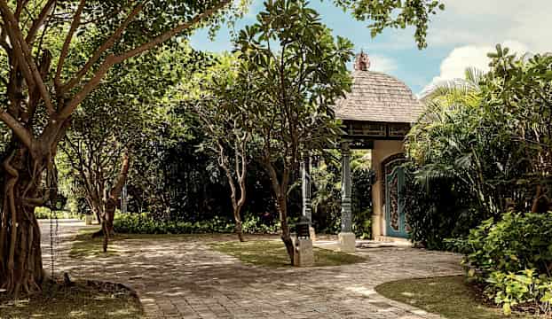 En väg inramad av grönska leder till en byggnad med ett dekorerat tak och dörr. (AI-genererad text)