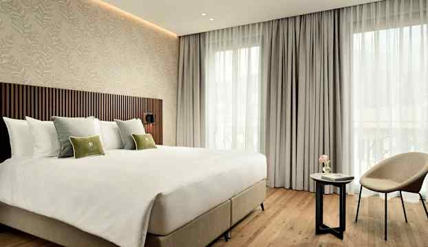 Ett modernt hotellrum med en stor säng, dekorativa kuddar, en fåtölj och ett mindre bord framför en fönstervägg med ljusa gardiner. (AI-genererad text)