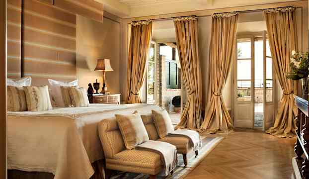 Ett elegant inrett sovrum med en stor säng, två fåtöljer och genomskinliga gardiner. En balkong syns genom glasdörrar. (AI-genererad text)